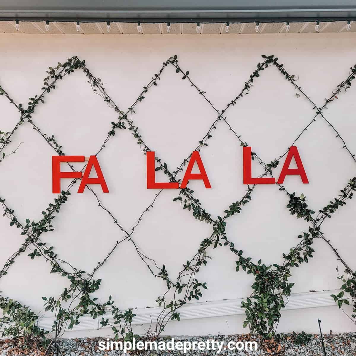 FA LA LA in large red letters on a white house wall behind a diamond lattice of lights beside a front door with a wreath